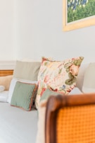 A cozy seating area with a collection of cushions, including a prominently placed large floral cushion with intricate designs. On the wall above is a framed painting with green foliage and blue flowers. The couch and cushions have muted tones, adding to the serene atmosphere.