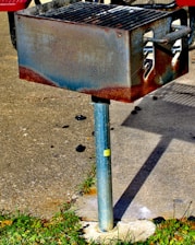 A rusty outdoor metal grill mounted on a metal pole, situated on a concrete surface with grass surrounding the base. Charcoal remnants are visible nearby.