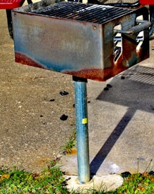 A rusty outdoor metal grill mounted on a metal pole, situated on a concrete surface with grass surrounding the base. Charcoal remnants are visible nearby.