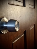 A close-up view of a metallic door knob fixed on a wooden door. The surface of the door shows visible grains and textures, indicating a natural wood finish. The lighting highlights the metallic sheen of the knob and the subtle patterns in the wood.