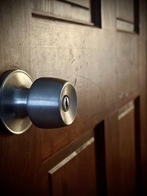 A close-up view of a metallic door knob fixed on a wooden door. The surface of the door shows visible grains and textures, indicating a natural wood finish. The lighting highlights the metallic sheen of the knob and the subtle patterns in the wood.