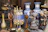 A collection of items including large decorative vases with blue and white floral patterns, various metal and ceramic ornaments, and statues. Among the statues are two children holding a red apple, and a figure carrying a basket on its head. The scene is cluttered with a variety of textures and colors.