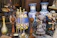 A collection of items including large decorative vases with blue and white floral patterns, various metal and ceramic ornaments, and statues. Among the statues are two children holding a red apple, and a figure carrying a basket on its head. The scene is cluttered with a variety of textures and colors.