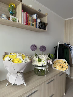 A wooden cabinet with various floral arrangements on top, including a bouquet of yellow and white flowers, a vase with white and purple flowers, and a bouquet of pale yellow roses. Above the cabinet, there is a shelf holding books, a plush toy, a cactus ornament, and decorative spheres. To the right, clothes are hanging on a rack.