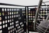 A balcony with a perforated metal panel railing featuring an array of circular cutouts. There is a hanging chair made of woven material with cushions, positioned to the right. The background is blurred, suggesting an outdoor setting.