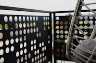 A balcony with a perforated metal panel railing featuring an array of circular cutouts. There is a hanging chair made of woven material with cushions, positioned to the right. The background is blurred, suggesting an outdoor setting.