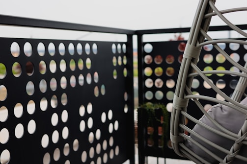 A balcony with a perforated metal panel railing featuring an array of circular cutouts. There is a hanging chair made of woven material with cushions, positioned to the right. The background is blurred, suggesting an outdoor setting.