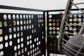A balcony with a perforated metal panel railing featuring an array of circular cutouts. There is a hanging chair made of woven material with cushions, positioned to the right. The background is blurred, suggesting an outdoor setting.