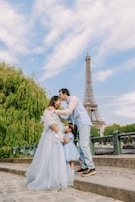 A family consisting of two adults and a child are standing outdoors near a large iconic tower. The adults are sharing a tender moment, and the child stands close to them, wearing a dress that matches the adult woman. Lush greenery and blue skies surround them, creating a picturesque setting.