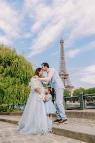A family consisting of two adults and a child are standing outdoors near a large iconic tower. The adults are sharing a tender moment, and the child stands close to them, wearing a dress that matches the adult woman. Lush greenery and blue skies surround them, creating a picturesque setting.