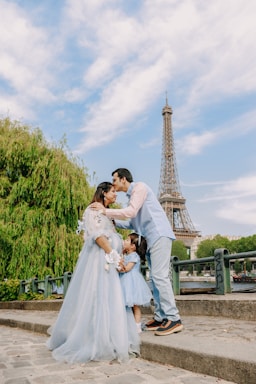 A family consisting of two adults and a child are standing outdoors near a large iconic tower. The adults are sharing a tender moment, and the child stands close to them, wearing a dress that matches the adult woman. Lush greenery and blue skies surround them, creating a picturesque setting.