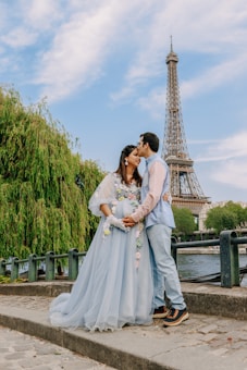 A couple stands embracing in front of the Eiffel Tower. The woman is wearing a light blue gown adorned with floral decorations, and the man is dressed in a light blue shirt with pants. They are surrounded by greenery and a river on a cobblestone path. The man gently kisses the woman's forehead.