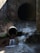 A concrete drainage pipe empties water into a stream, creating small waterfalls. The scene is set against a rough, weathered wall with some patches of greenery. The water flow is steady, and the surrounding area is damp and earthy, suggesting a functional but neglected infrastructure.