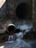 A concrete drainage pipe empties water into a stream, creating small waterfalls. The scene is set against a rough, weathered wall with some patches of greenery. The water flow is steady, and the surrounding area is damp and earthy, suggesting a functional but neglected infrastructure.