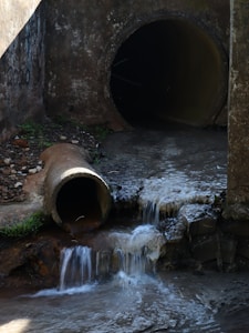A concrete drainage pipe empties water into a stream, creating small waterfalls. The scene is set against a rough, weathered wall with some patches of greenery. The water flow is steady, and the surrounding area is damp and earthy, suggesting a functional but neglected infrastructure.