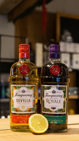 Two bottles of Tanqueray gin are displayed on a wooden surface, one labeled Sevilla Distilled Gin with vibrant orange and citrus-themed packaging, and the other labeled Dark Berry Royale with dark purple and berry-themed packaging. A lemon half is placed in front of the bottles. The background is a blurred indoor setting with shelves and various products.