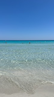 Crystal clear turquoise waters stretch out towards the horizon, under a vast clear blue sky. Gentle waves lap onto the sandy shore, creating a serene and tranquil atmosphere. A few people are visible in the distance, enjoying the calmness of the open sea.