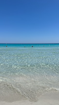 Crystal clear turquoise waters stretch out towards the horizon, under a vast clear blue sky. Gentle waves lap onto the sandy shore, creating a serene and tranquil atmosphere. A few people are visible in the distance, enjoying the calmness of the open sea.