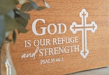 A wooden plaque featuring the phrase 'God is our refuge and strength' from Psalm 46:1 is displayed, with a decorative cross design beside the text. A blurred green plant is visible on the left in a glass vase.