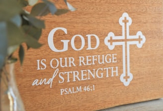 A wooden plaque featuring the phrase 'God is our refuge and strength' from Psalm 46:1 is displayed, with a decorative cross design beside the text. A blurred green plant is visible on the left in a glass vase.