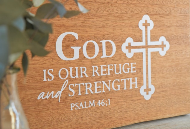 A wooden plaque featuring the phrase 'God is our refuge and strength' from Psalm 46:1 is displayed, with a decorative cross design beside the text. A blurred green plant is visible on the left in a glass vase.