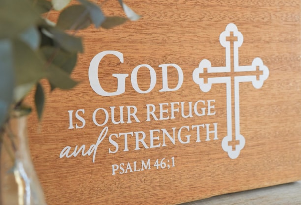 A wooden plaque featuring the phrase 'God is our refuge and strength' from Psalm 46:1 is displayed, with a decorative cross design beside the text. A blurred green plant is visible on the left in a glass vase.