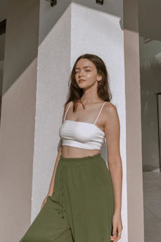 A stylish young woman wearing a breezy summer outfit, standing against a soft beige wall with sunlight casting gentle shadows.