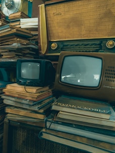 A vintage setup featuring old televisions and a radio placed on top of stacks of books. The televisions have small screens and the radio has a classic wooden design. The overall setting suggests a nostalgic, retro ambiance with elements of clutter and age.