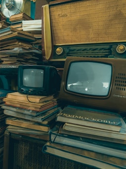A vintage setup featuring old televisions and a radio placed on top of stacks of books. The televisions have small screens and the radio has a classic wooden design. The overall setting suggests a nostalgic, retro ambiance with elements of clutter and age.