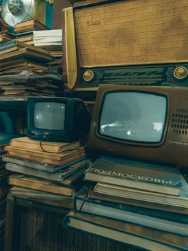 A vintage setup featuring old televisions and a radio placed on top of stacks of books. The televisions have small screens and the radio has a classic wooden design. The overall setting suggests a nostalgic, retro ambiance with elements of clutter and age.