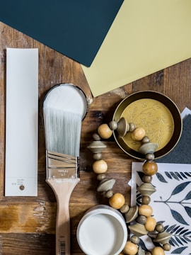 A variety of painting and decorative items are arranged on a wooden surface. There are paint samples, a paintbrush with white paint, a white paint can, wooden beads on a string, and a decorative golden container with a floral design. The background includes a paint swatch in dark green and light yellow colors.