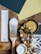 A variety of painting and decorative items are arranged on a wooden surface. There are paint samples, a paintbrush with white paint, a white paint can, wooden beads on a string, and a decorative golden container with a floral design. The background includes a paint swatch in dark green and light yellow colors.