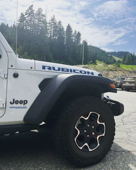 A white Rubicon Jeep Wrangler is parked outdoors on a paved surface. The angle captures the front left side of the vehicle, highlighting the wheel, fender, and hood with badges visible. In the background, there is a scenic view of lush green pine trees and hills under a partly cloudy sky.