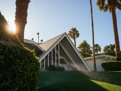 A modern house with a distinctive triangular roof is surrounded by lush greenery and palm trees. Sunlight filters through the foliage, casting a warm glow over the scene. Water sprays from a lawn sprinkler, indicating a moment of care for the vibrant, well-maintained garden.