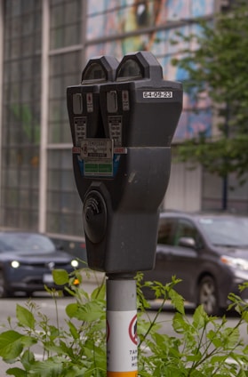 A close-up view of a parking meter situated on a sidewalk. The parking meter is mounted on a pole with various informational labels, including a temporary parking permit notice. In the background, a blurred image of a busy street is visible, with cars and greenery partially in focus. The surroundings are urban, with a multicolored mural or glass panels on a building across the street.