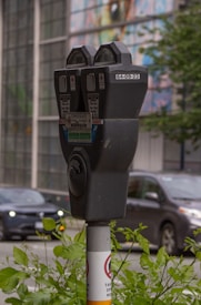 A close-up view of a parking meter situated on a sidewalk. The parking meter is mounted on a pole with various informational labels, including a temporary parking permit notice. In the background, a blurred image of a busy street is visible, with cars and greenery partially in focus. The surroundings are urban, with a multicolored mural or glass panels on a building across the street.