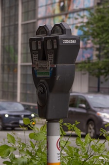 A close-up view of a parking meter situated on a sidewalk. The parking meter is mounted on a pole with various informational labels, including a temporary parking permit notice. In the background, a blurred image of a busy street is visible, with cars and greenery partially in focus. The surroundings are urban, with a multicolored mural or glass panels on a building across the street.