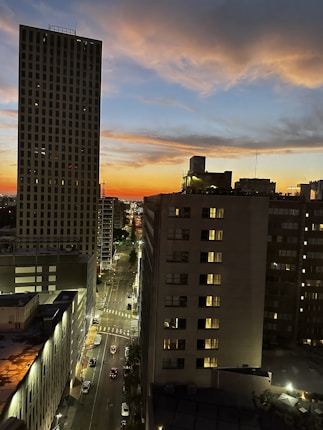 A cityscape at dusk with tall buildings lining a street, illuminated by streetlights and the warm glow from windows. The sky showcases a gradient of colors from blue to orange, with clouds scattered across.
