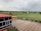 A school courtyard features a red-roofed building with glass windows on the left and a yellow school bus parked on a paved area. Beyond the fence, open green fields extend to distant houses and trees under a cloudy sky.