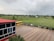 A school courtyard features a red-roofed building with glass windows on the left and a yellow school bus parked on a paved area. Beyond the fence, open green fields extend to distant houses and trees under a cloudy sky.