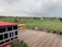 A school courtyard features a red-roofed building with glass windows on the left and a yellow school bus parked on a paved area. Beyond the fence, open green fields extend to distant houses and trees under a cloudy sky.