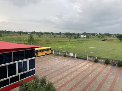 A school courtyard features a red-roofed building with glass windows on the left and a yellow school bus parked on a paved area. Beyond the fence, open green fields extend to distant houses and trees under a cloudy sky.