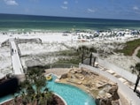 A view of a sandy beach with a clear, blue ocean in the background. Many people are scattered along the beach, under umbrellas and engaging in recreational activities. A boardwalk leads to the beach area, and a swimming pool with a waterfall feature is surrounded by palm trees and a sunbathing area in the foreground.