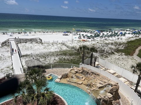 A view of a sandy beach with a clear, blue ocean in the background. Many people are scattered along the beach, under umbrellas and engaging in recreational activities. A boardwalk leads to the beach area, and a swimming pool with a waterfall feature is surrounded by palm trees and a sunbathing area in the foreground.