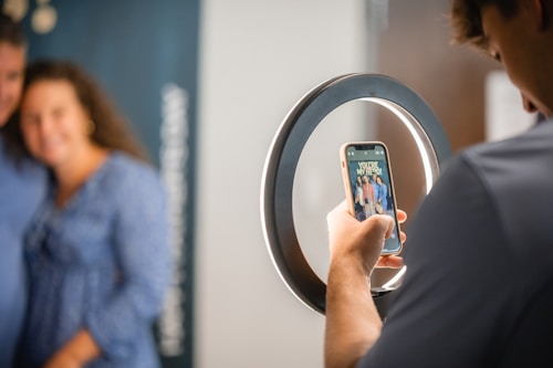 A person is taking a photo of a blurred couple using a smartphone through a ring light. The smartphone displays the message 'YOU'RE MY HERO!' with an image of a group of people. The background is soft and out of focus, creating a professional and intimate setting.