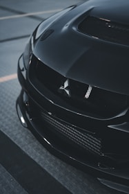 Front view of a sleek black car with a Mitsubishi emblem on the grille. The car features carbon fiber details and has a sporty and aggressive look, captured in a dimly lit parking area.