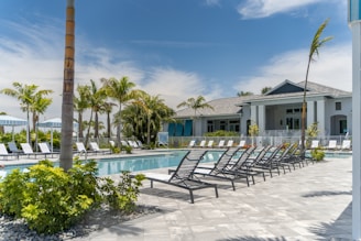 A luxurious outdoor swimming pool area surrounded by numerous reclining chairs, palm trees, and a modern building in the background. The sky is clear with a few clouds visible. The setting conveys a tranquil and inviting atmosphere, ideal for relaxation and leisure.