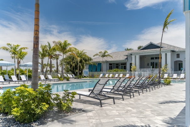 A luxurious outdoor swimming pool area surrounded by numerous reclining chairs, palm trees, and a modern building in the background. The sky is clear with a few clouds visible. The setting conveys a tranquil and inviting atmosphere, ideal for relaxation and leisure.