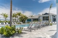 A luxurious outdoor swimming pool area surrounded by numerous reclining chairs, palm trees, and a modern building in the background. The sky is clear with a few clouds visible. The setting conveys a tranquil and inviting atmosphere, ideal for relaxation and leisure.