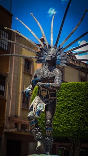 A detailed and ornate statue of a person wearing traditional attire and an impressive headdress stands prominently. The headdress is adorned with long feathers, and the person holds decorative items in each hand. The statue features intricate patterns and embellishments in tones of blue and gold with a backdrop of a green tree and a clear blue sky.
